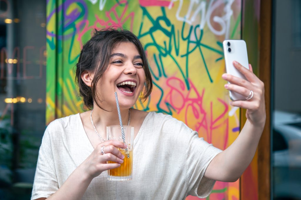 mulher tira selfie para o instagram enquanto toma um drink em frente a uma parede com grafite colorido em bar