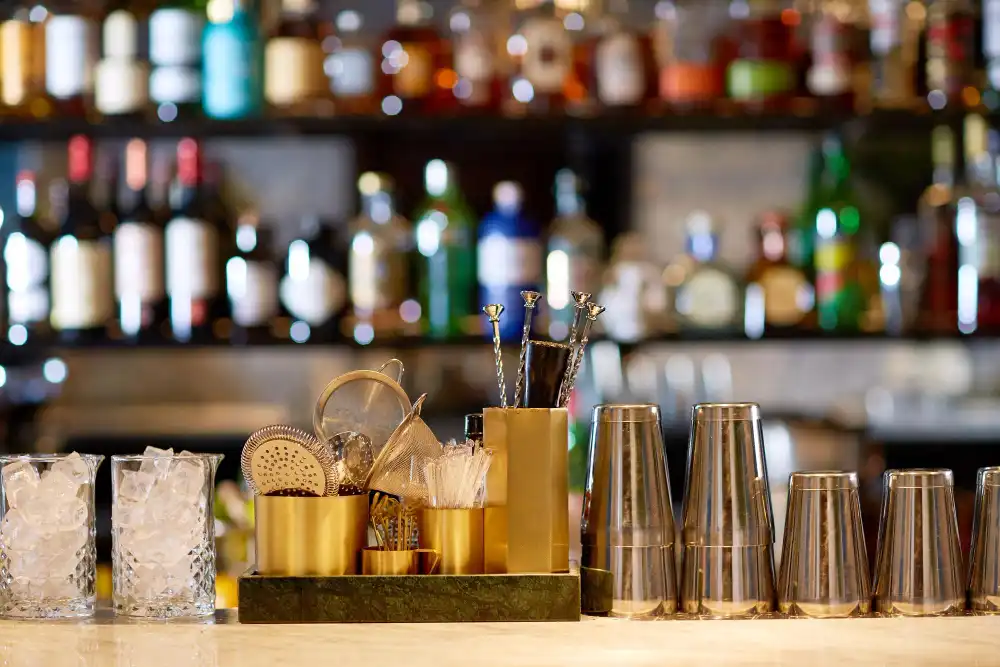 Equipamentos para bar incluindo copos, utensílios e acessórios dispostos em uma bancada, com garrafas ao fundo, ideal para profissionais da coquetelaria.