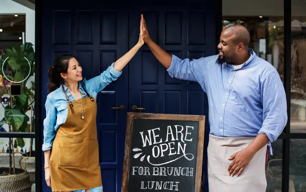 Casal animado inaugurando um novo restaurante, mostrando a placa de aberto com entusiasmo. Exemplificando o momento de abrir um restaurante.