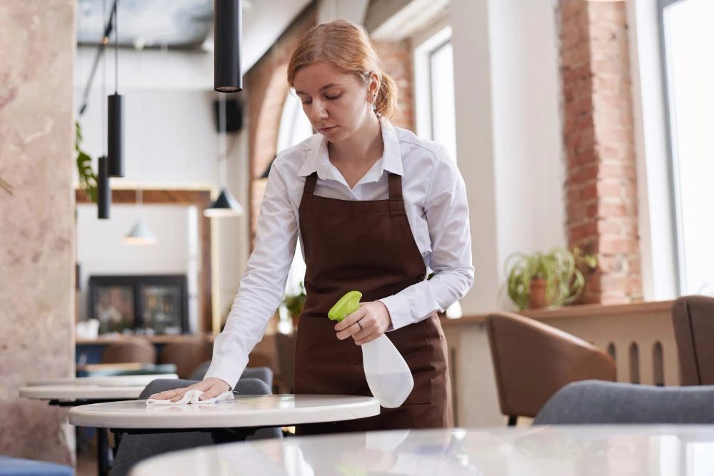 	
Funcionária de restaurante realizando a limpeza de uma mesa com spray desinfetante e pano, em referência aos equipamentos de limpeza.