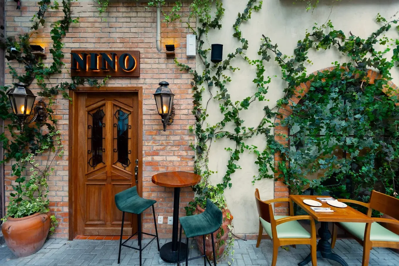 Fachada do Nino, com letreiro iluminado, hera nas paredes de tijolo e mesas na calçada