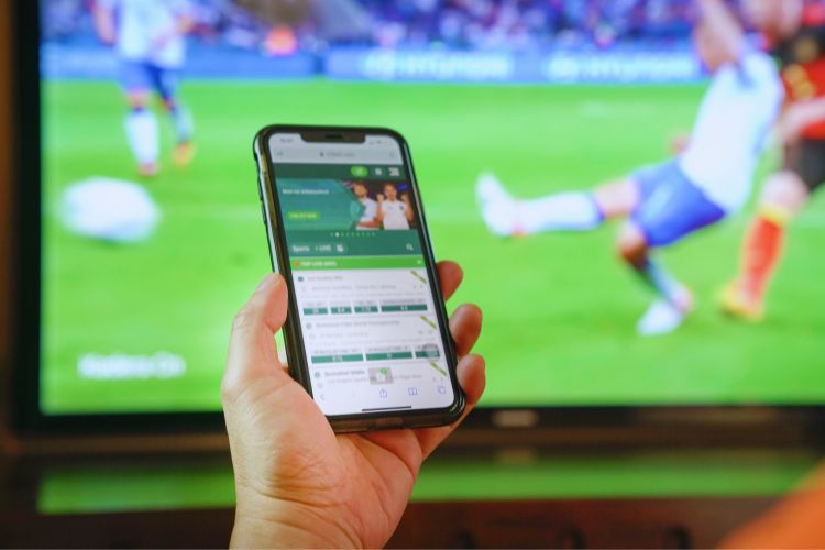 Mão segurando smartphone mostrando tela de aplicativo de bets, com uma TV passando futebol ao fundo